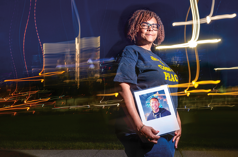 Cheryl Graham-Seay stands holding a photo of her son, Jarell Christopher Seay, at night, with a city skyline behind her.